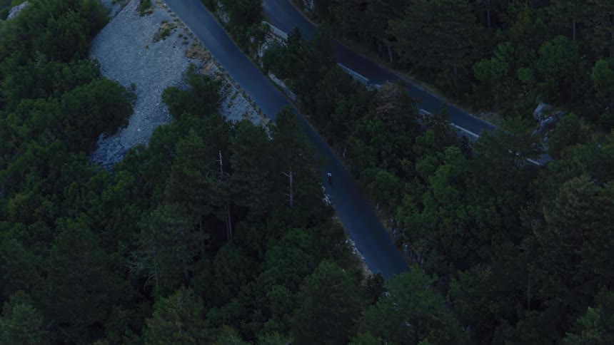 High angle aerial shot follows a group of cyclists descending a winding switchback road through a dense, dark mountain forest