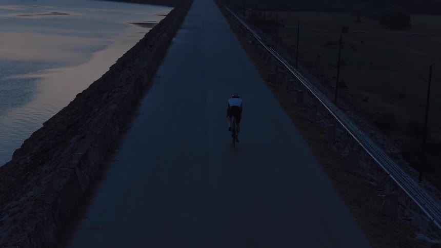 Aerial view of a solitary cyclist riding on a long, empty road across a dam at twilight with mountains in the distance