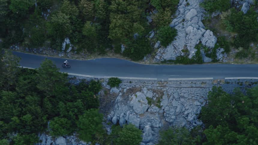 Aerial top-down shot of a cyclist riding along a winding mountain road surrounded by trees and rocks