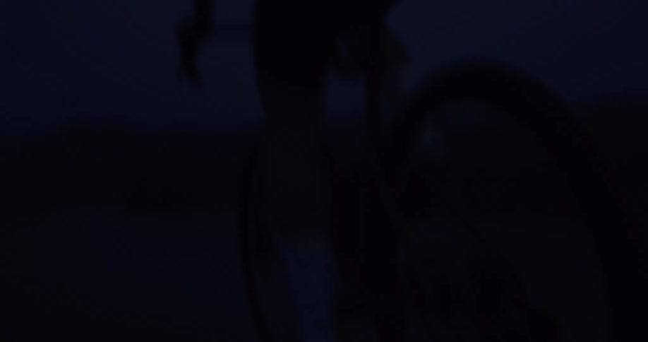 A low-angle close-up shot captures the silhouette of a cyclist's legs and a spinning wheel as they ride at night