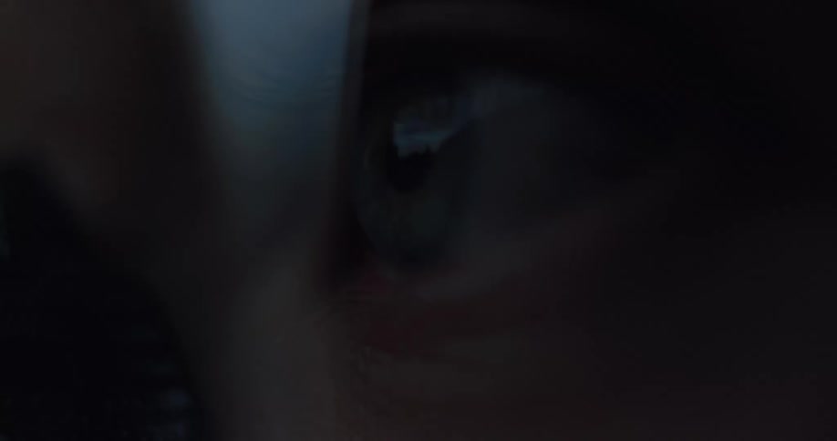 An extreme close-up of a human eye watching intently in a dark, moody setting, with a screen reflected in the pupil