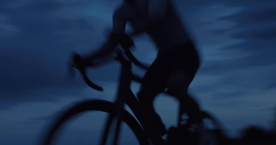 A silhouetted cyclist rides intensely toward the camera on a dark road at dusk