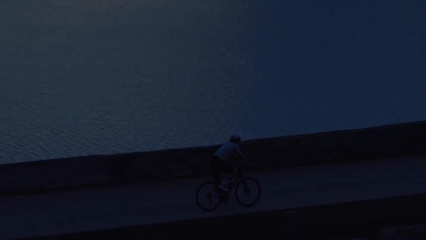High-angle shot follows a cyclist riding along a tranquil waterfront in the deep blue twilight