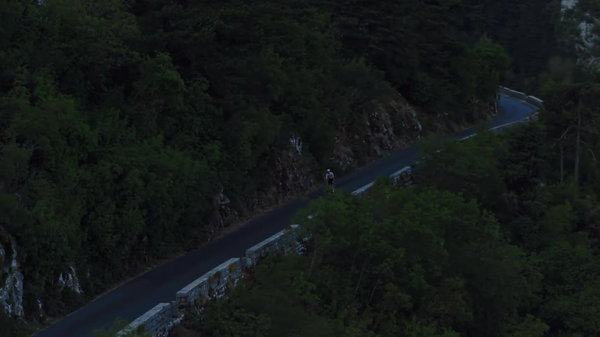 An aerial view follows a lone cyclist navigating a winding road through a dense, dark mountain forest at dusk