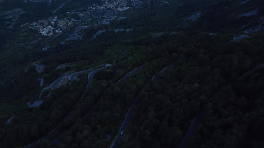 Eerie twilight aerial of a serpentine road winding through a dark mountain forest towards a distant, glowing village