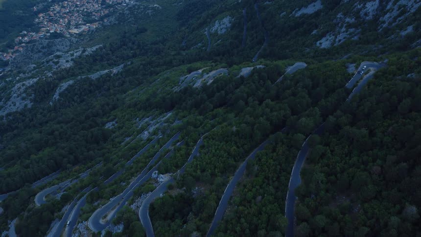 A dramatic aerial view reveals a winding serpentine road with hairpin turns cutting through a dense forest on a mountainside at dusk