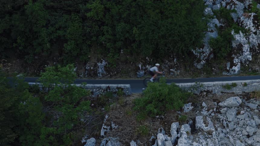 Aerial tracking shot of a cyclist riding a winding road on a rocky, forested mountainside