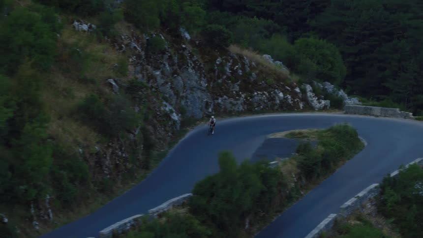An aerial view shows a lone cyclist climbing a winding mountain pass at dusk