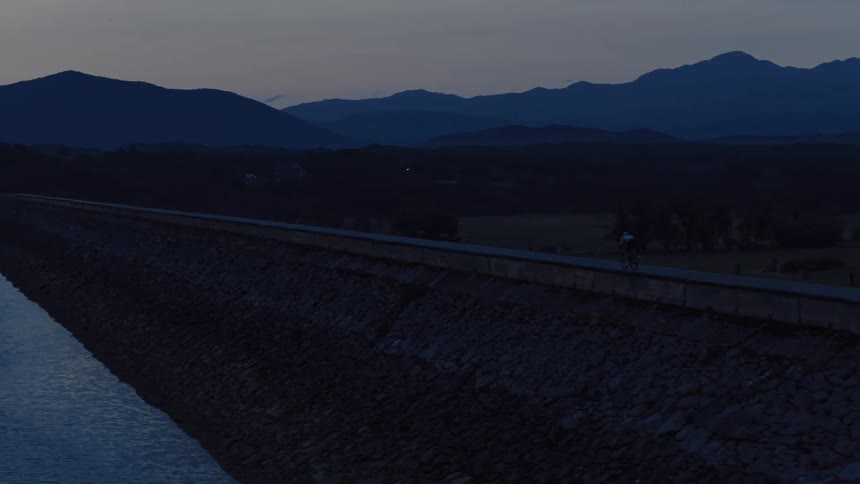 Aerial view of a lone cyclist riding along a quiet dam road next to a calm lake at twilight, with mountains silhouetted against the fading light