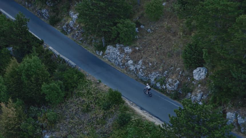 An aerial top-down view captures a lone cyclist ascending a winding mountain road through a lush green forest