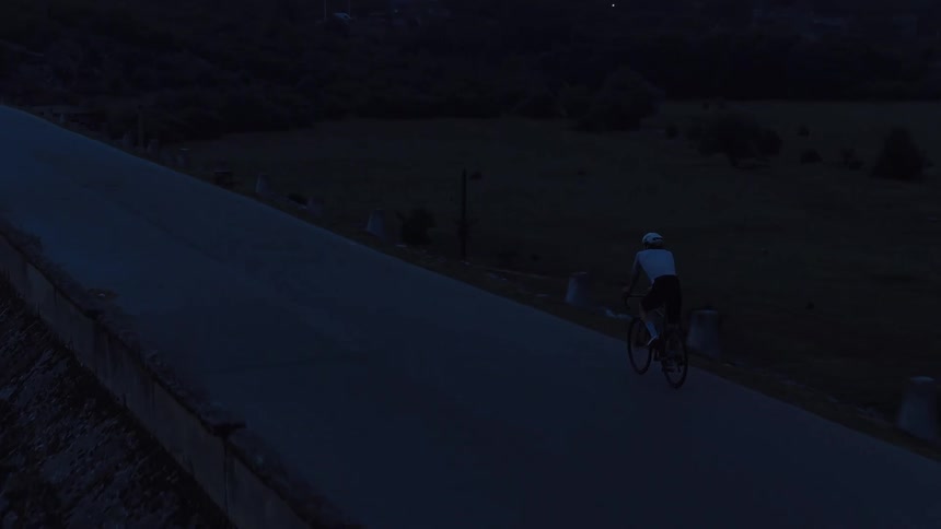 An aerial view follows a lone cyclist riding a mountain road at dusk