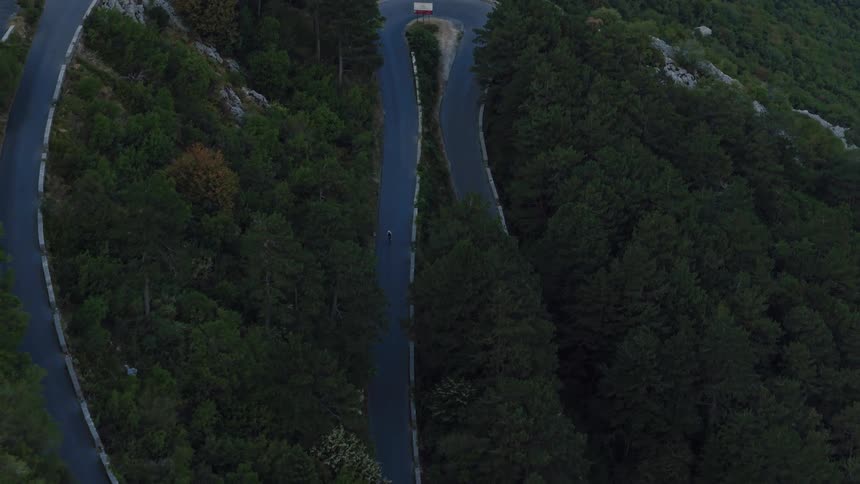 A lone cyclist descends a serpentine road winding through a dense mountain forest in this dramatic aerial shot