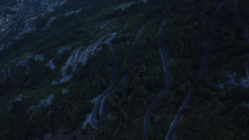 An aerial view captures the dramatic switchbacks of a mountain road through a dark forest at twilight