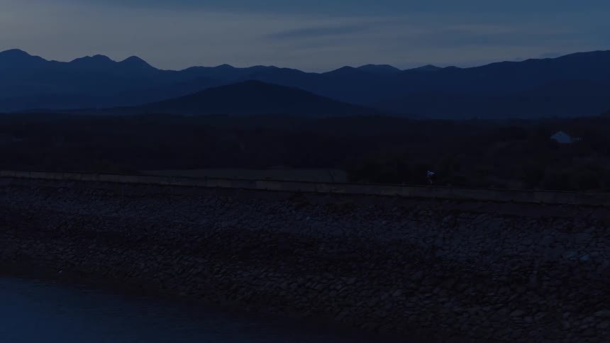 A drone follows a lone cyclist riding along a dam at twilight with a majestic mountain range in the background