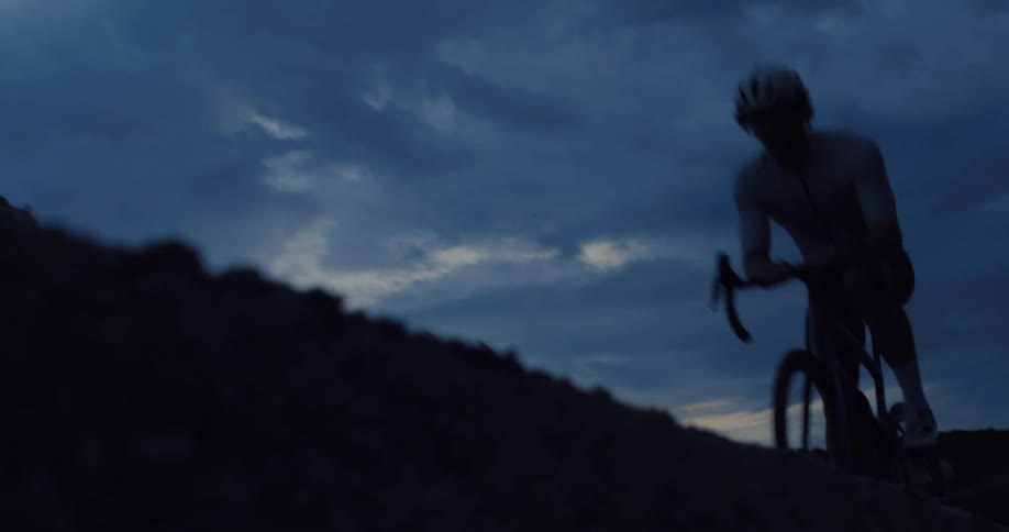 A determined cyclist races past the camera in slow motion while climbing a hill at dusk