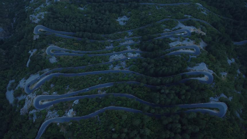 An aerial top-down view shows cars navigating the numerous hairpin turns of a winding serpentine road up a forested mountain at dusk