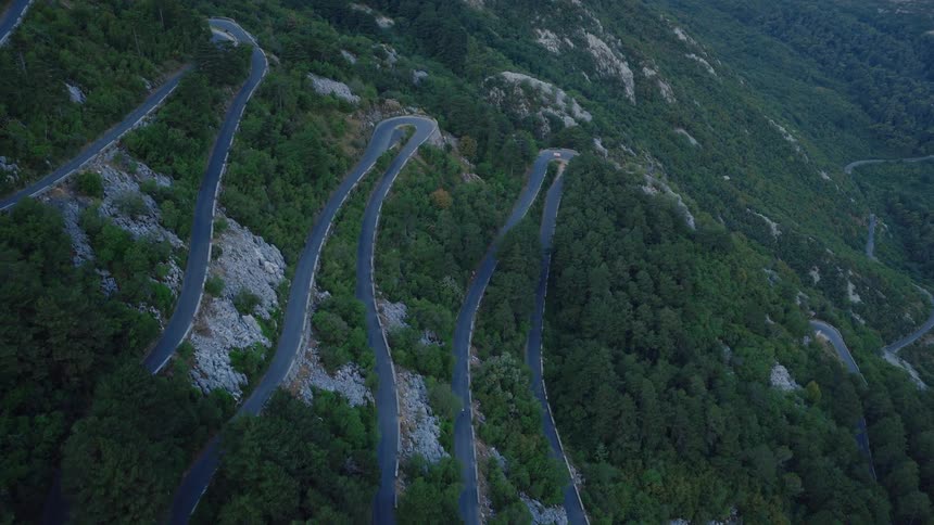 Epic aerial view of a serpentine mountain road winding through a dense green forest with dramatic hairpin turns