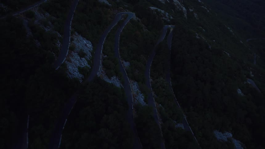 A slow aerial reveal of a lone car with headlights on driving up a dark, winding mountain pass at dusk