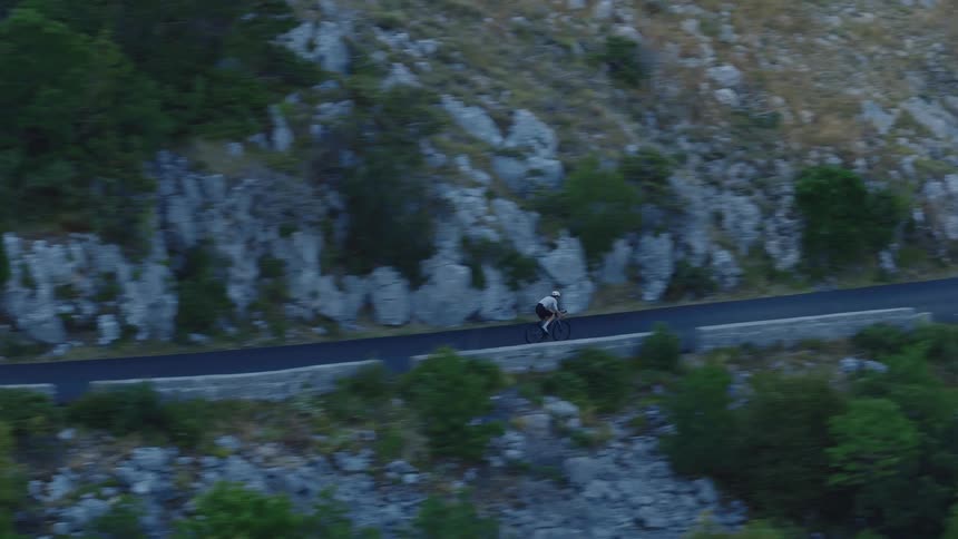 An aerial tracking shot follows a lone cyclist riding along a paved road carved into a steep, rocky mountainside with sparse green trees