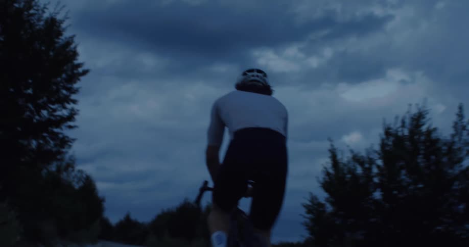 Low angle rear view of a cyclist riding on a rural road at dusk under a dark and cloudy sky