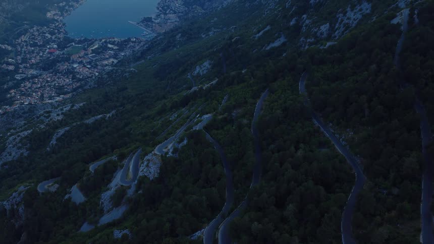 An aerial view pulls back to reveal a winding serpentine road up a mountainside overlooking a coastal town and bay at dusk