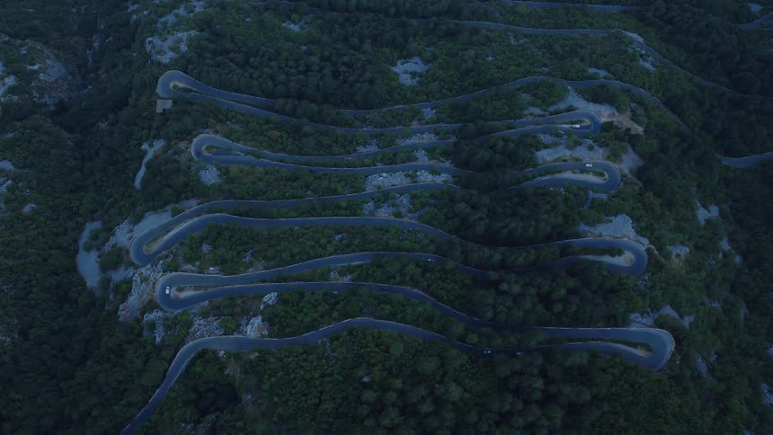 An aerial top-down view shows cars navigating the serpentine switchbacks of a mountain road surrounded by a dense forest at dusk