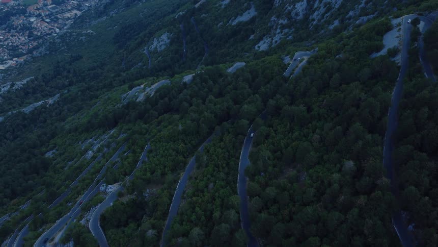 A high aerial view follows cars navigating a winding serpentine road with multiple switchbacks up a lush, tree-covered mountainside