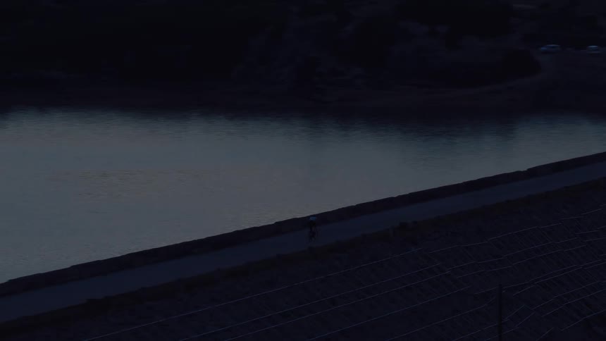 Aerial footage captures a solitary cyclist pedaling along a remote path beside a tranquil reservoir during the quiet hours of dusk