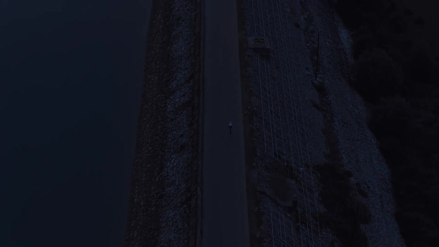 An aerial view follows a lone cyclist riding on a path along a reservoir dam during the moody blue hour of dusk