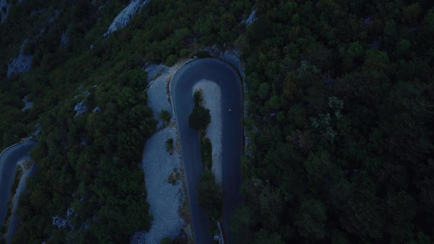 A lone cyclist navigates a tight hairpin turn on a winding mountain pass surrounded by a dense forest at dusk, as seen from a top-down aerial view