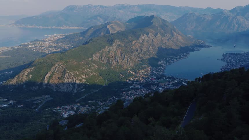 Epic aerial shot over forested mountains looking down on a tranquil bay and coastal town at sunrise