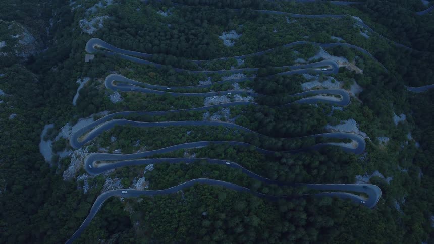 An aerial top-down view shows cars navigating the sharp turns of a serpentine road on a forested mountainside at dusk