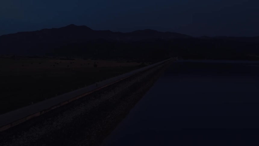 An aerial view shows a lone cyclist riding along a quiet road on top of a dam next to a reservoir at twilight