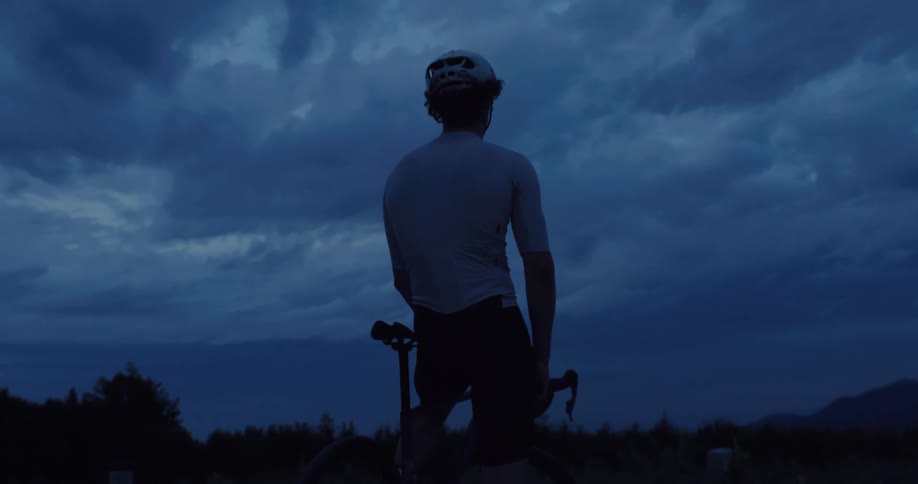 A cinematic slow-motion shot shows a male cyclist resting on his bike at dusk, silhouetted against a dramatic and moody mountain sky