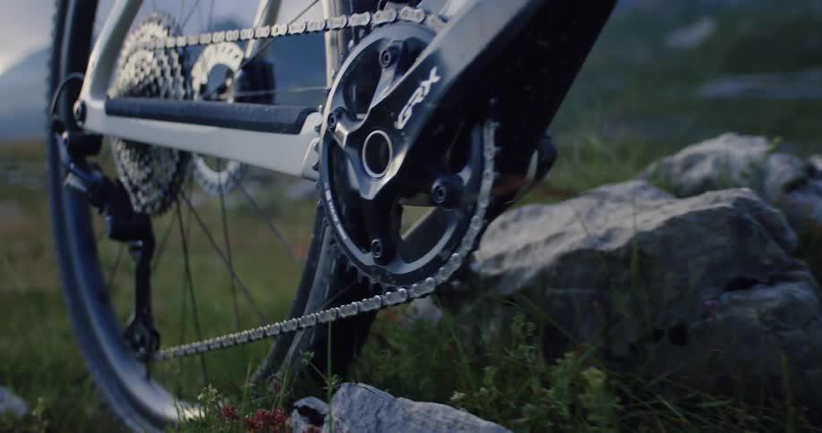 A close-up shot of a gravel bike's drivetrain, including the chain, gears, and crankset, resting on a rocky, grassy trail in the mountains