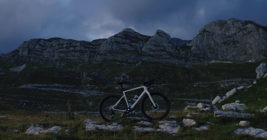 A lone white bicycle stands against a dramatic backdrop of rugged mountains under a cloudy twilight sky