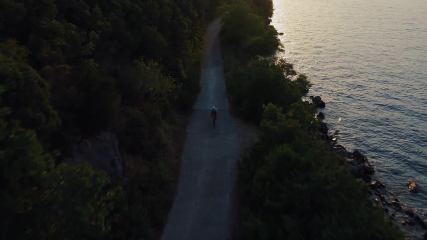 An aerial view captures a lone cyclist riding along a scenic coastal path next to the ocean at sunset