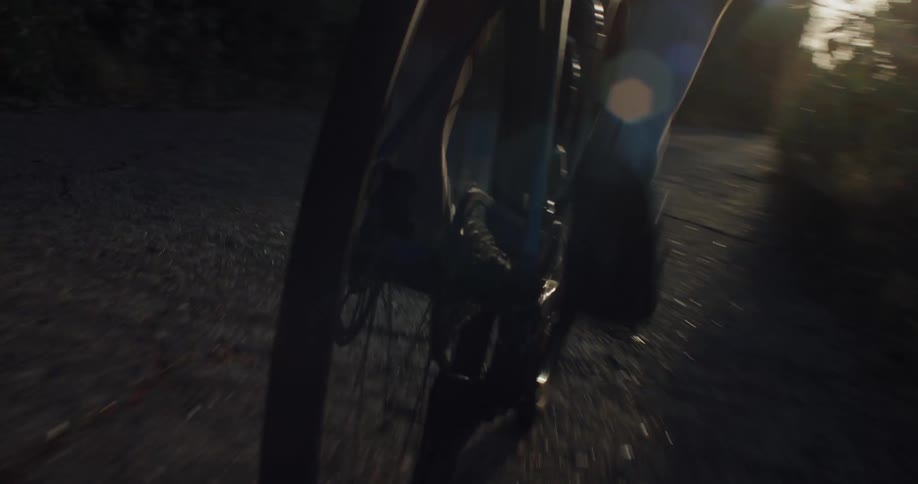 A close-up low-angle tracking shot of a cyclist's legs and feet pedaling on a road bike with the golden hour sun creating a lens flare