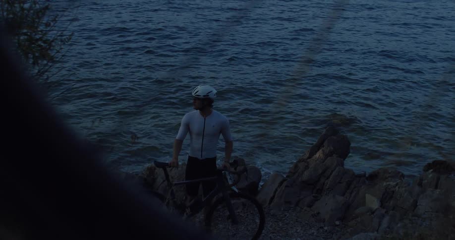 A pensive cyclist stands with his bike on a rocky shore at dusk, looking out over the dark water