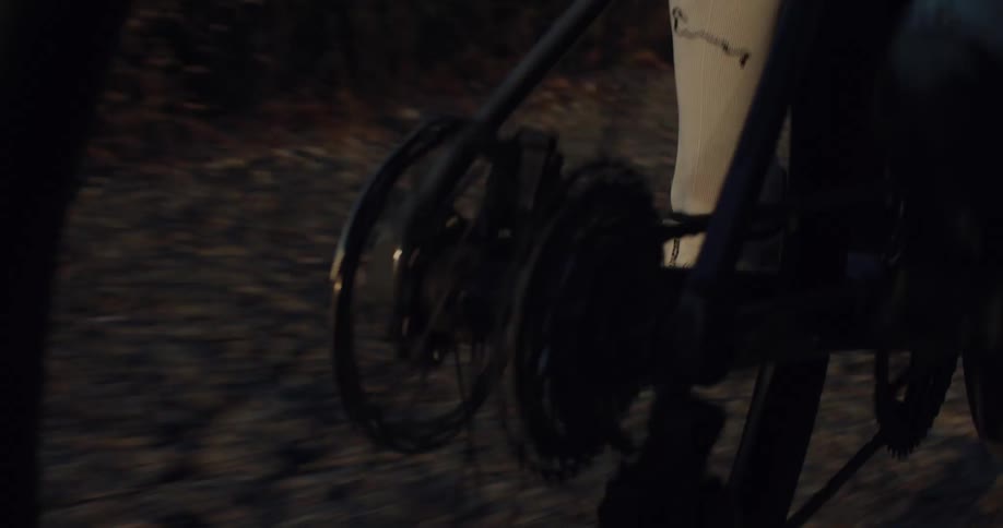 Close-up tracking shot of a bicycle's rear cassette and wheel spinning as a cyclist pedals along a road at dusk
