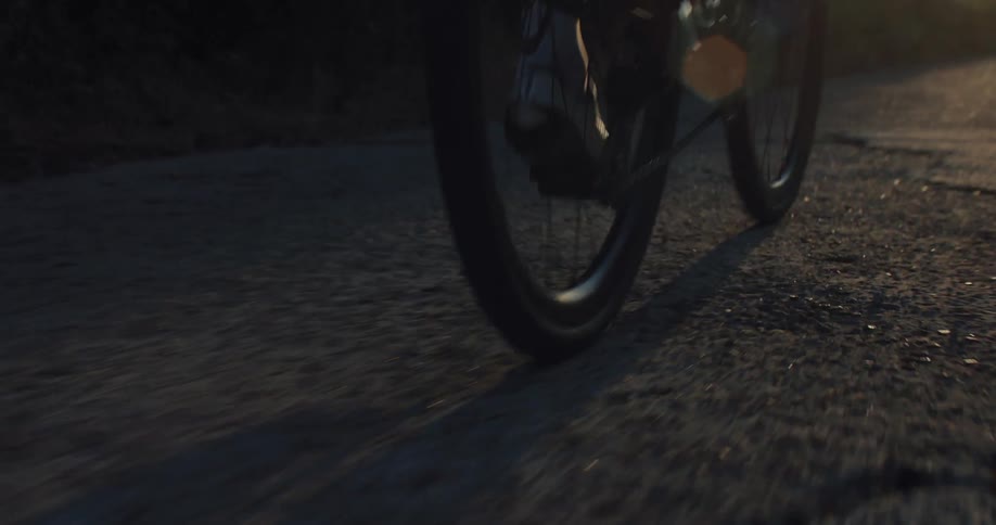 A slow-motion close-up from behind shows a cyclist's feet pedaling a road bike on an asphalt road during a beautiful sunset