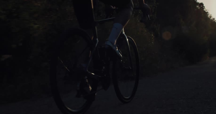 Cinematic slow motion tracking shot of a cyclist pedaling a road bike down a country lane at sunset