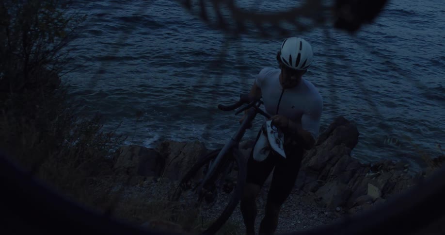 A cyclist in athletic gear carries his bike up a rocky hill next to the water at dusk