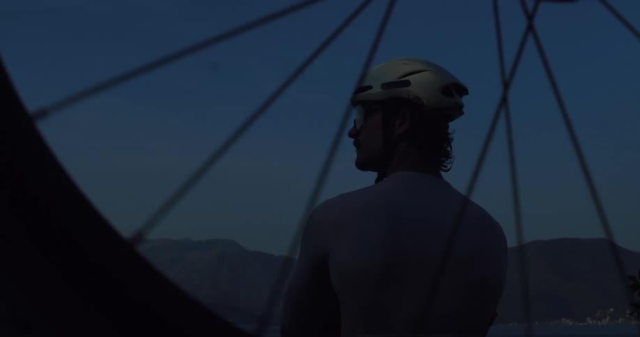 Through the spokes of a bicycle wheel, a cyclist takes a pensive break and enjoys the tranquil mountain and sea view at twilight