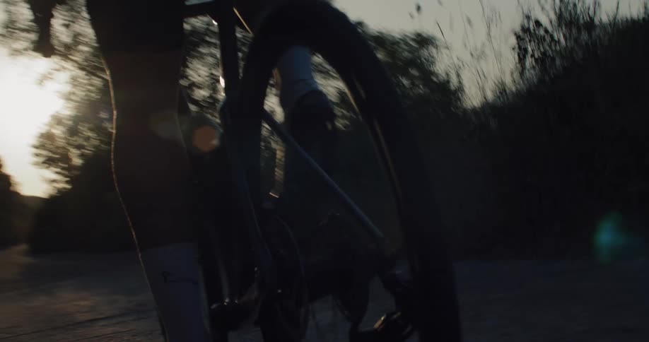Low angle close-up of a cyclist's legs and rear wheel as they pedal quickly down a paved path into the sunset