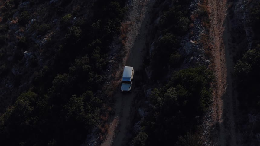 Top-down aerial view slowly rises above a white off-road vehicle on a remote mountain trail overlooking the deep blue sea