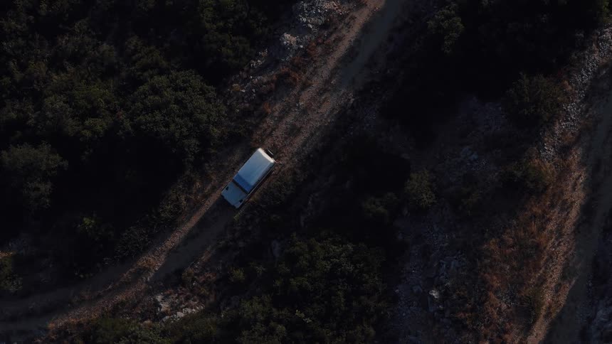 Aerial top-down shot of a white SUV driving on a remote dirt road through a shadowy, forested mountainside