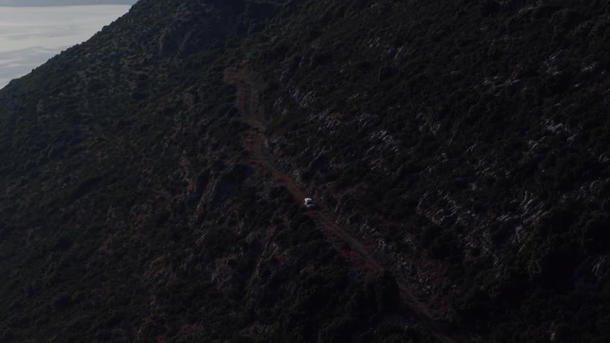 An aerial view captures a lone white vehicle driving along a winding dirt road on a rugged mountainside at dusk