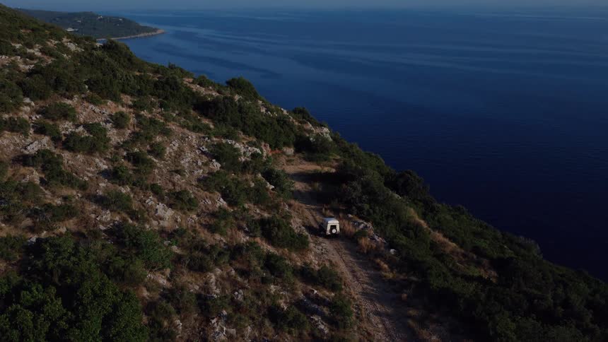 Aerial reveal of a white off-road vehicle parked on a scenic coastal mountain road overlooking the deep blue sea