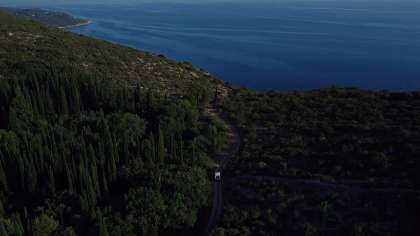 A campervan drives down a dirt road through a dense forest along the beautiful coastline, as seen from a high aerial perspective